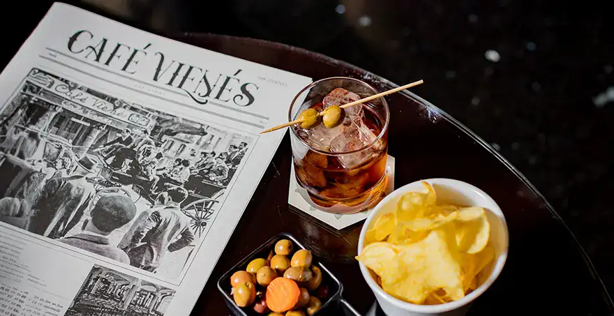 Peri&oacute;dico, vaso con bebida, aceitunas y papas sobre una mesa en un caf&eacute;.