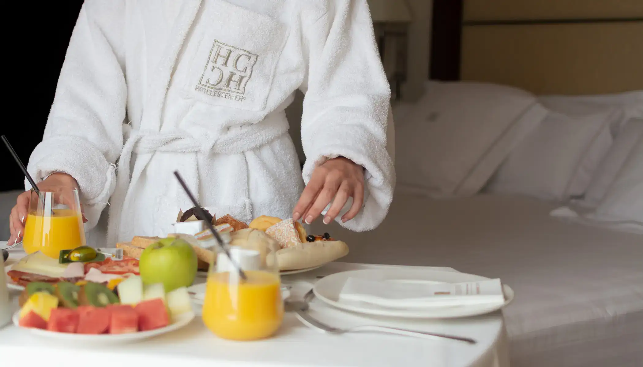 Persona en bata con desayuno sobre la cama, incluye jugo y fruta.
