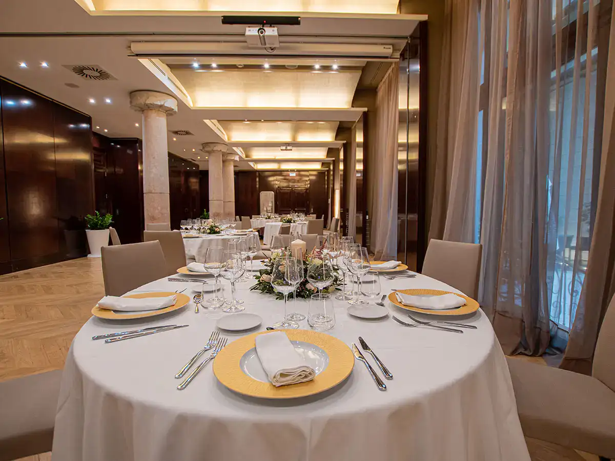 Mesa elegante en un sal&oacute;n, preparada para un evento formal con vajilla y copas.