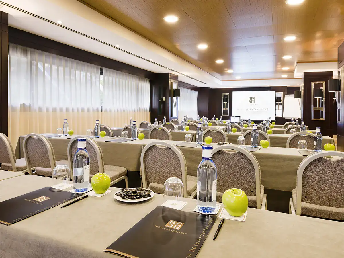 Sala de conferencias con mesas, sillas, botellas de agua y manzanas verdes.