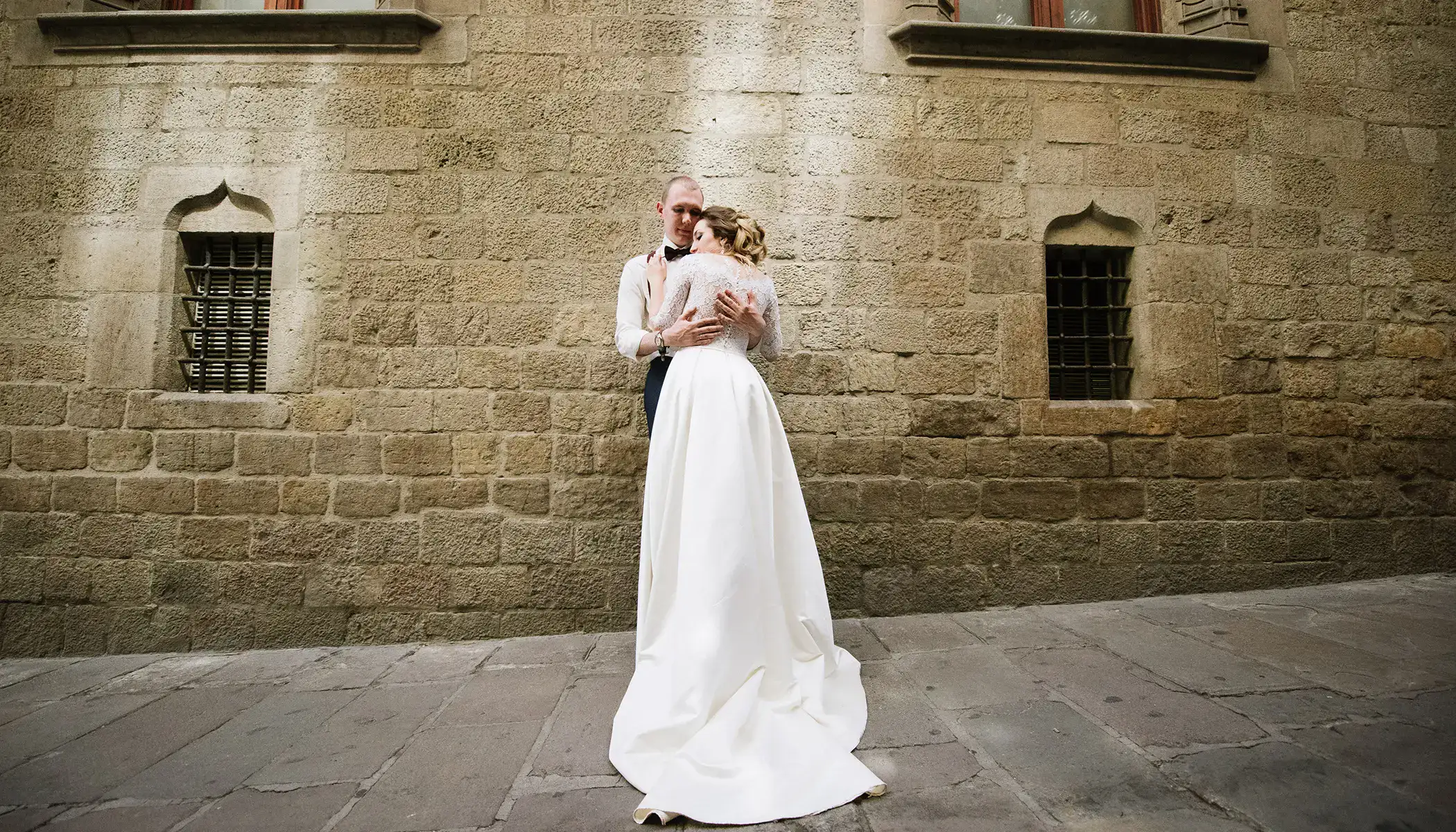 Pareja abraz&aacute;ndose frente a un muro de piedra antigua.