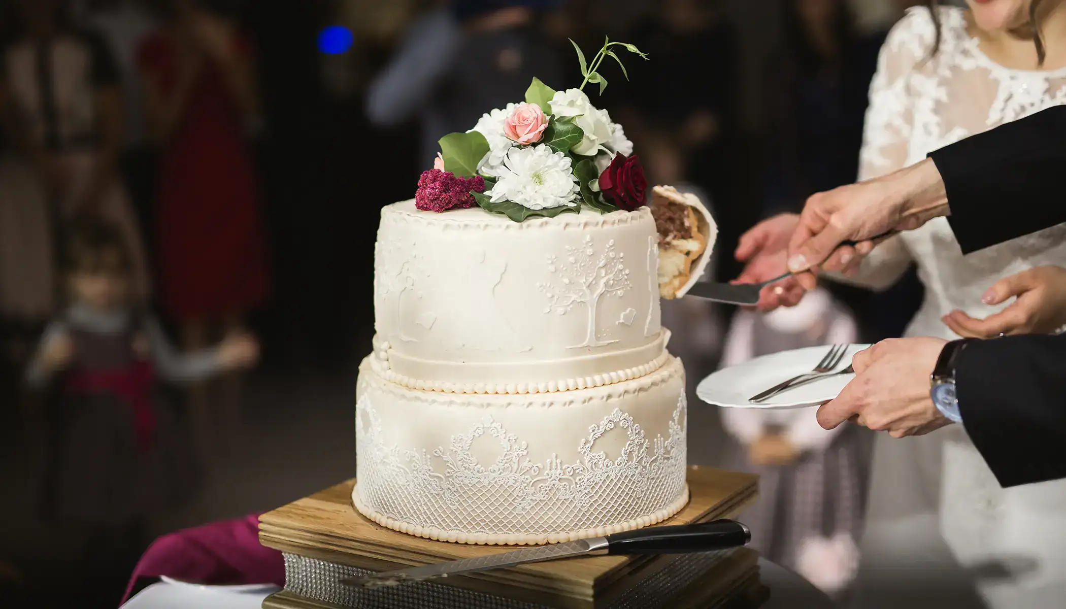 Pastel de boda blanco siendo cortado, decorado con flores y detalles en encaje.