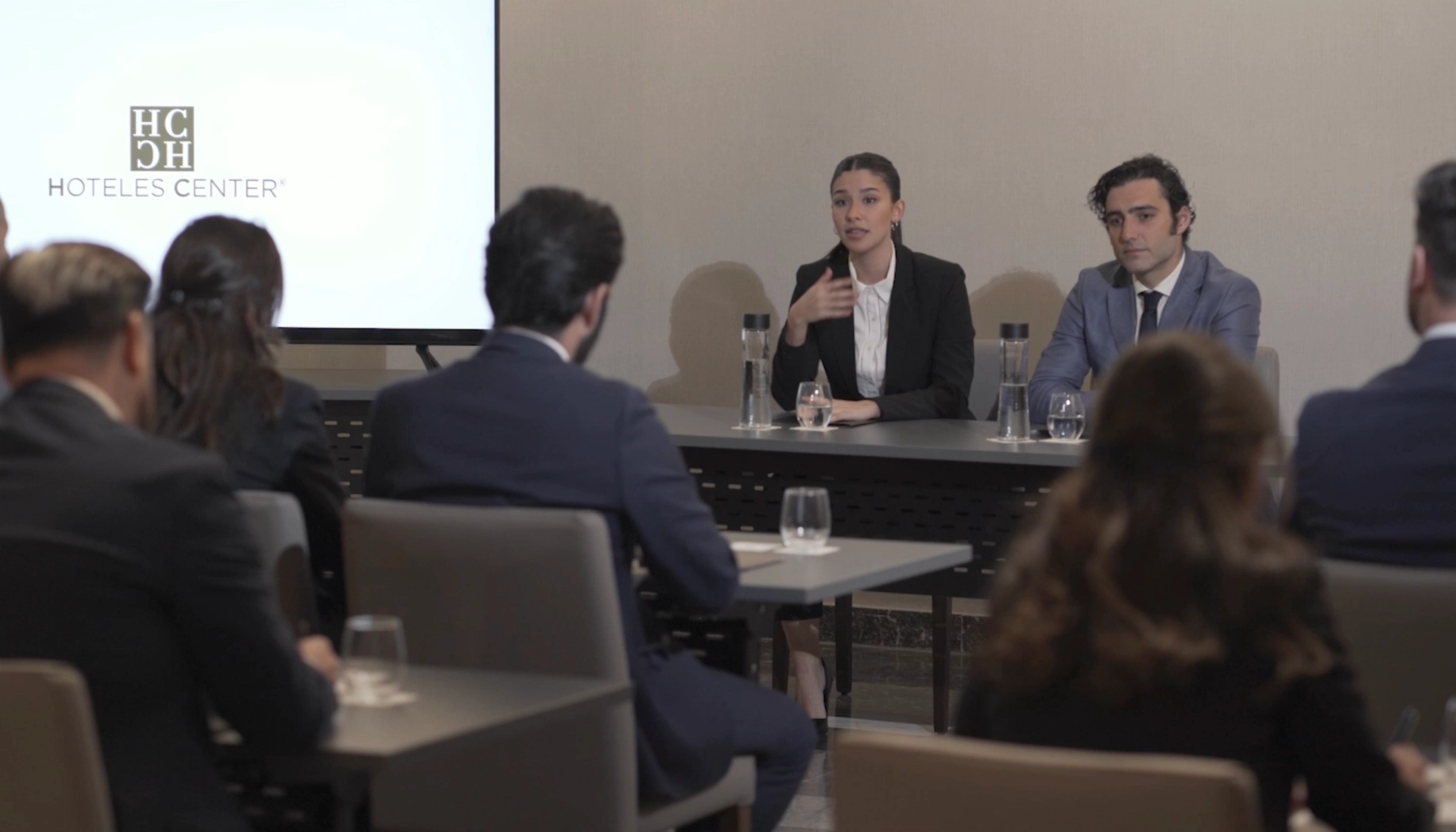 Personas en una reuni&oacute;n formal, hablando desde una mesa. Pantalla al fondo.