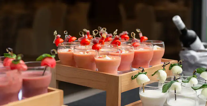 Vasos de gazpacho y tapas en una mesa; botella de vino al fondo.