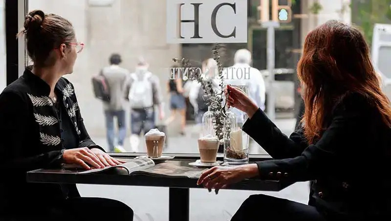 Dos mujeres conversan en una cafeter&iacute;a con bebidas y revistas sobre la mesa.