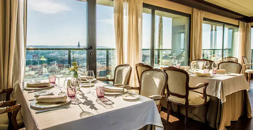 Restaurante con vista panor&aacute;mica de la ciudad, mesas elegantemente puestas.