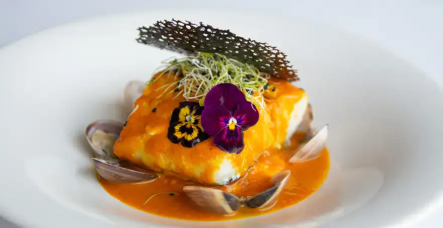 Plato gourmet con pescado en salsa naranja, flores comestibles y almejas.
