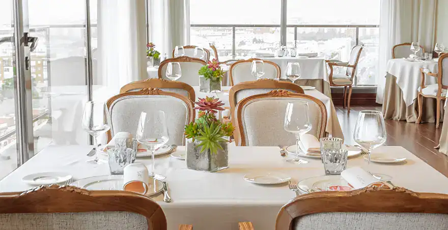 Restaurante elegante con mesas blancas y copas de vino junto a una ventana grande.