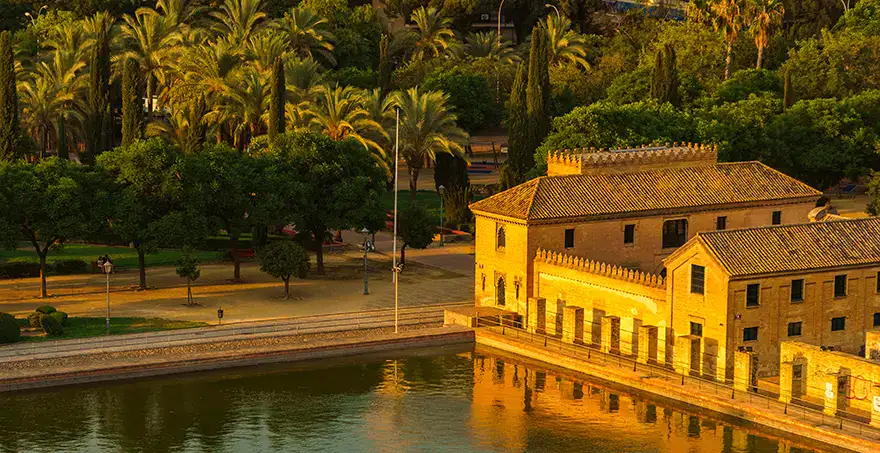 Edificio hist&oacute;rico junto a un r&iacute;o rodeado de &aacute;rboles y palmeras al atardecer.