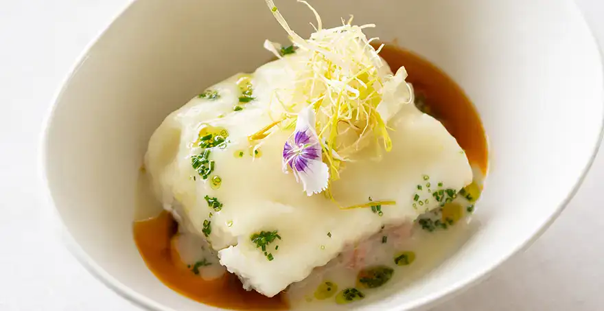 Plato de pescado con salsa, decorado con flores y hierbas en un bol blanco.