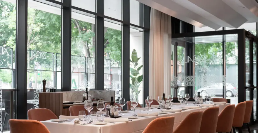 Restaurante elegante con mesas dispuestas, copas y vista a la calle arbolada.