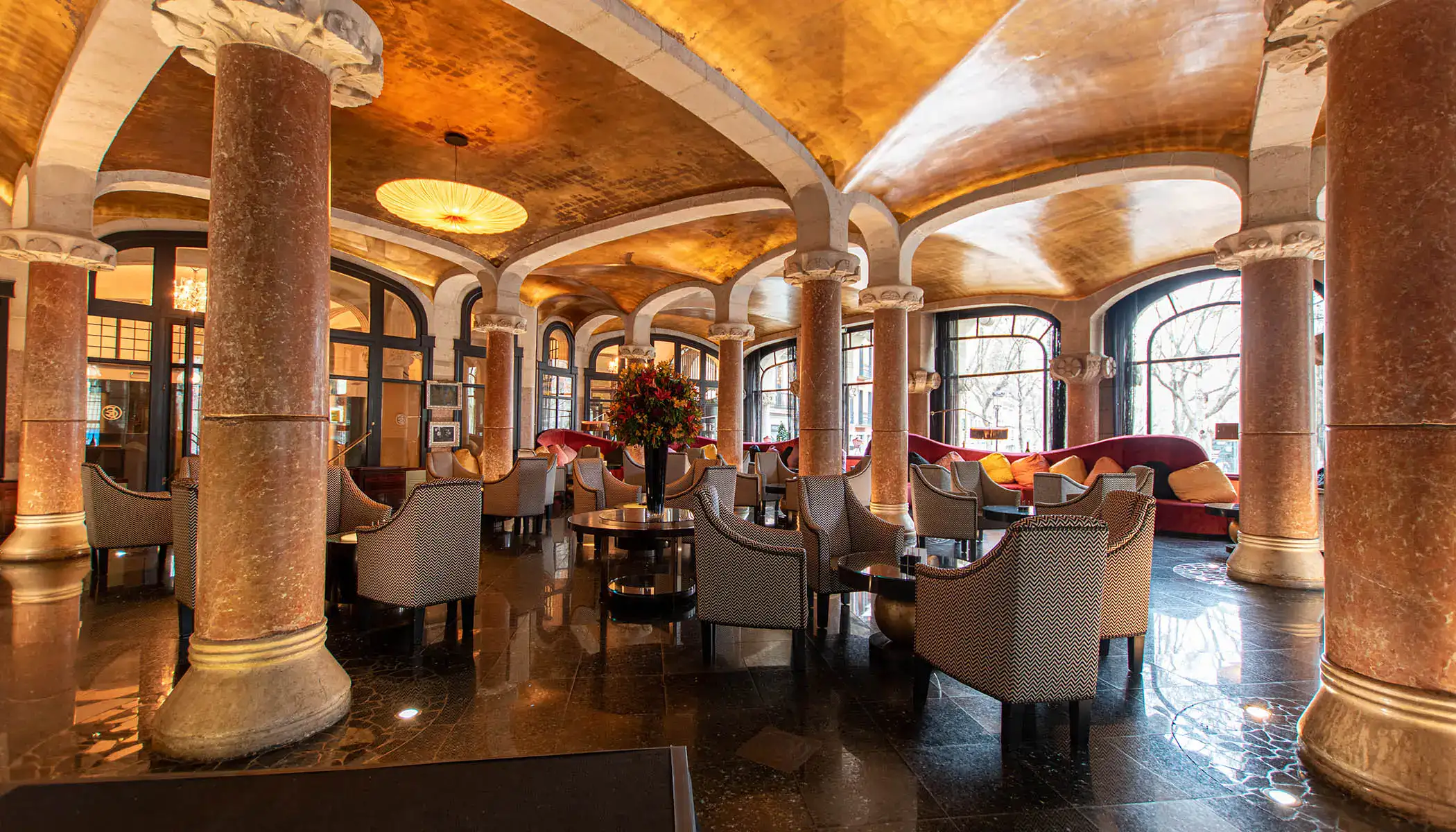 Elegante sal&oacute;n con columnas y techo dorado, sillas grises y mesa central.