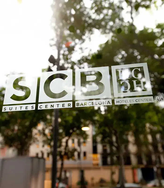 Logotipo de "Suites Center Barcelona" en un vidrio con fondo de &aacute;rboles.