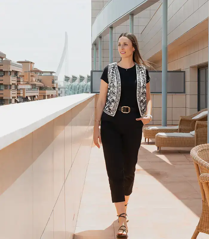 Mujer caminando en una terraza soleada con edificios y estructura moderna al fondo.