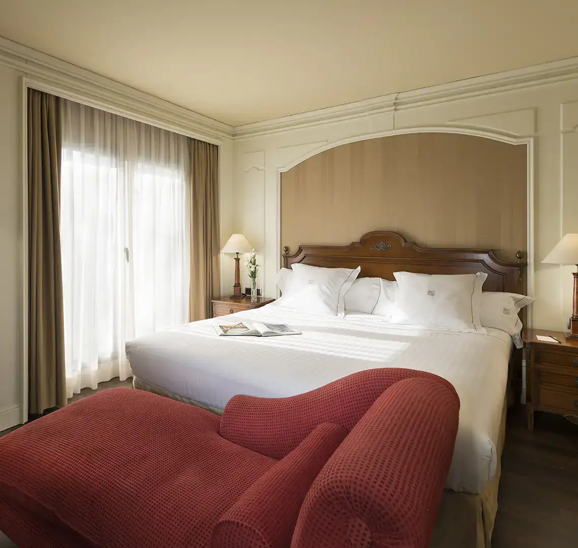 Habitaci&oacute;n elegante con cama grande, sill&oacute;n rojo y l&aacute;mpara de mesa.