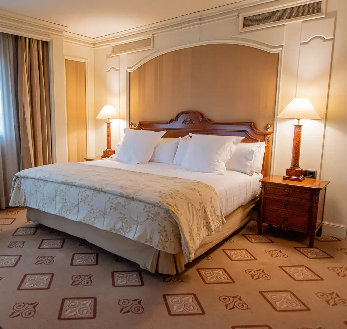 Cama doble con cabecero de madera, l&aacute;mparas encendidas y alfombra decorada.