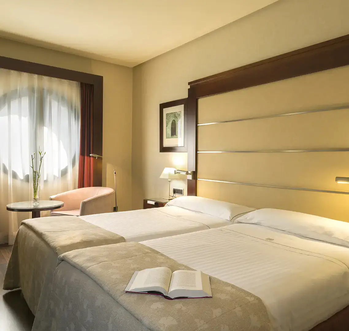 Habitaci&oacute;n de hotel con dos camas, una silla y un libro abierto.