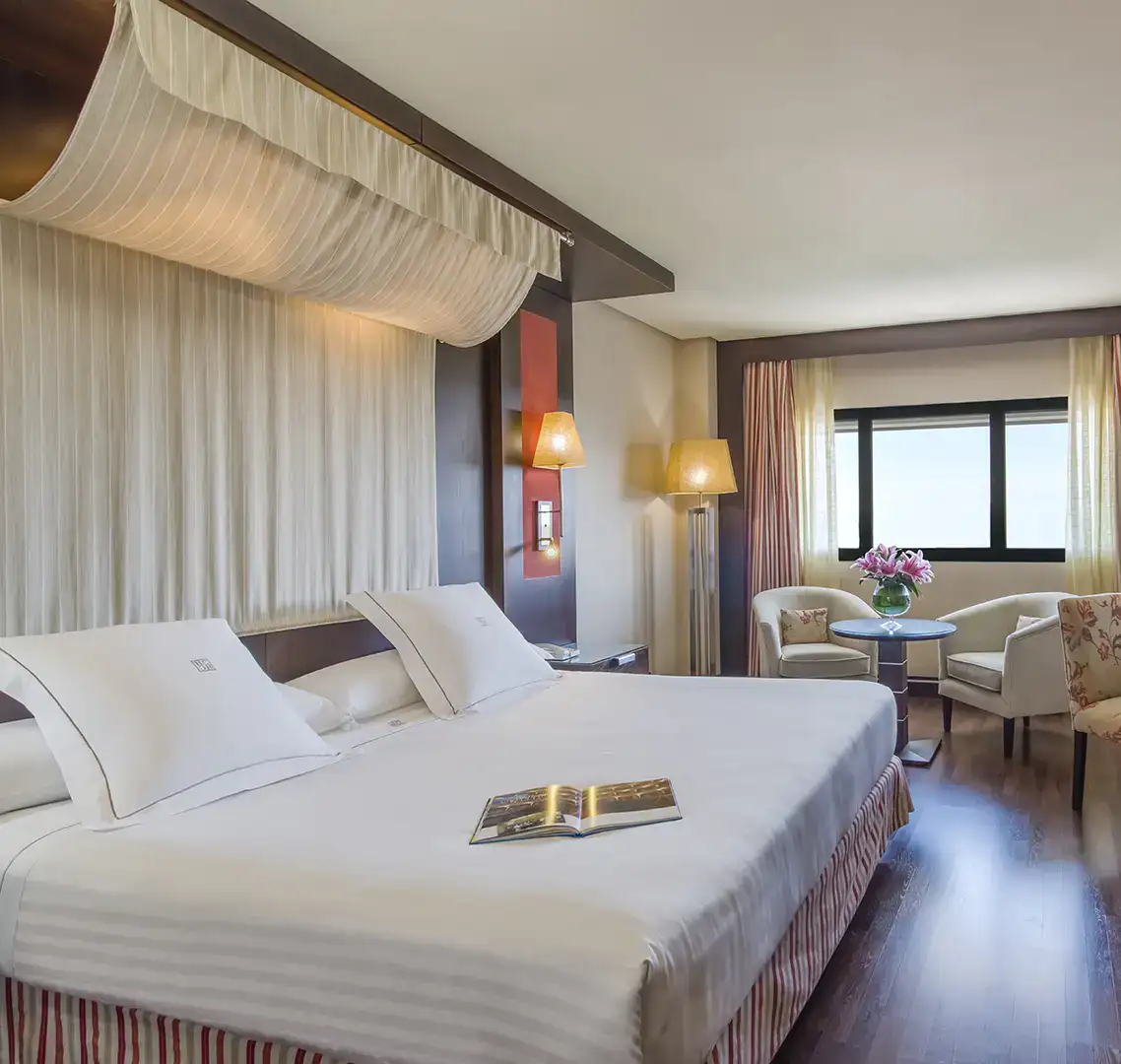 Habitaci&oacute;n de hotel elegante con cama grande y &aacute;rea de descanso junto a la ventana.