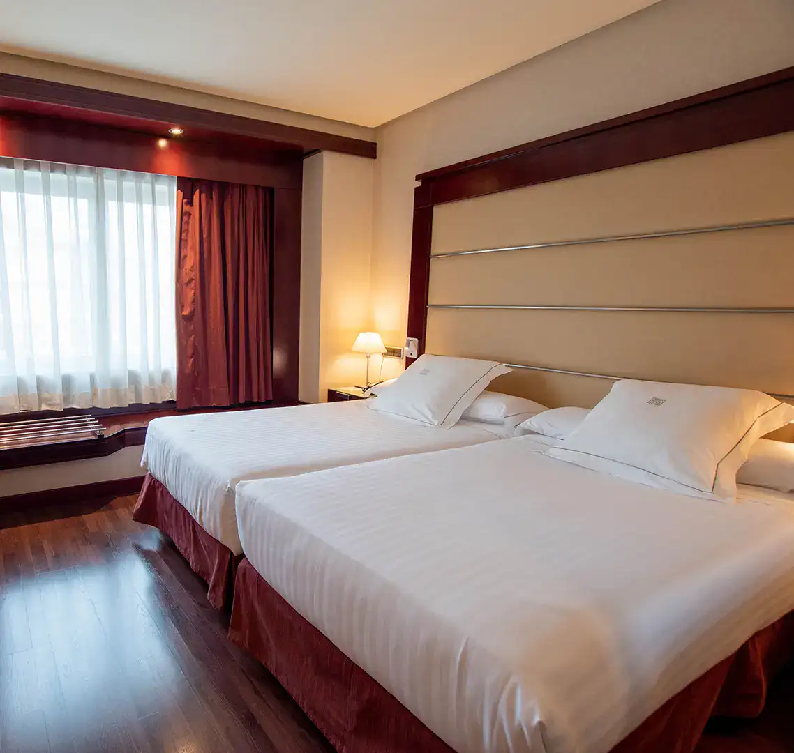 Habitaci&oacute;n de hotel con dos camas, ventana y luz tenue de l&aacute;mpara.