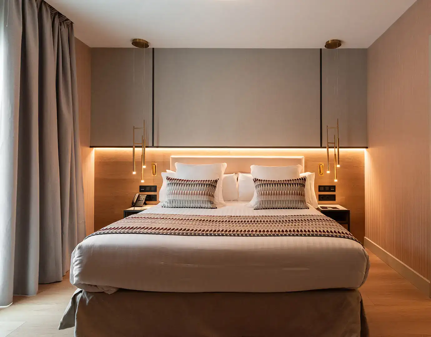 Cama doble con iluminaci&oacute;n moderna y cortinas en un dormitorio elegante.