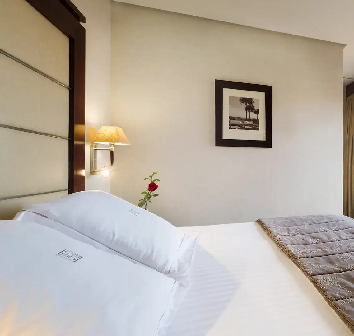 Cama con s&aacute;banas blancas y rosa roja en la mesita de noche. Cuadro en la pared.