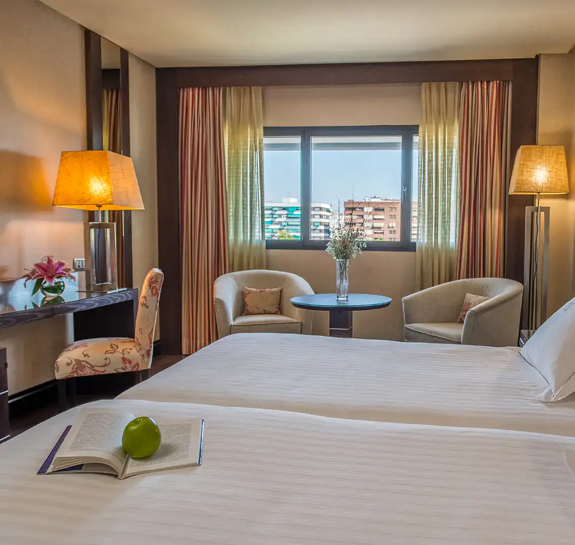 Habitaci&oacute;n de hotel luminosa con cama, sillones y vista a la ciudad.