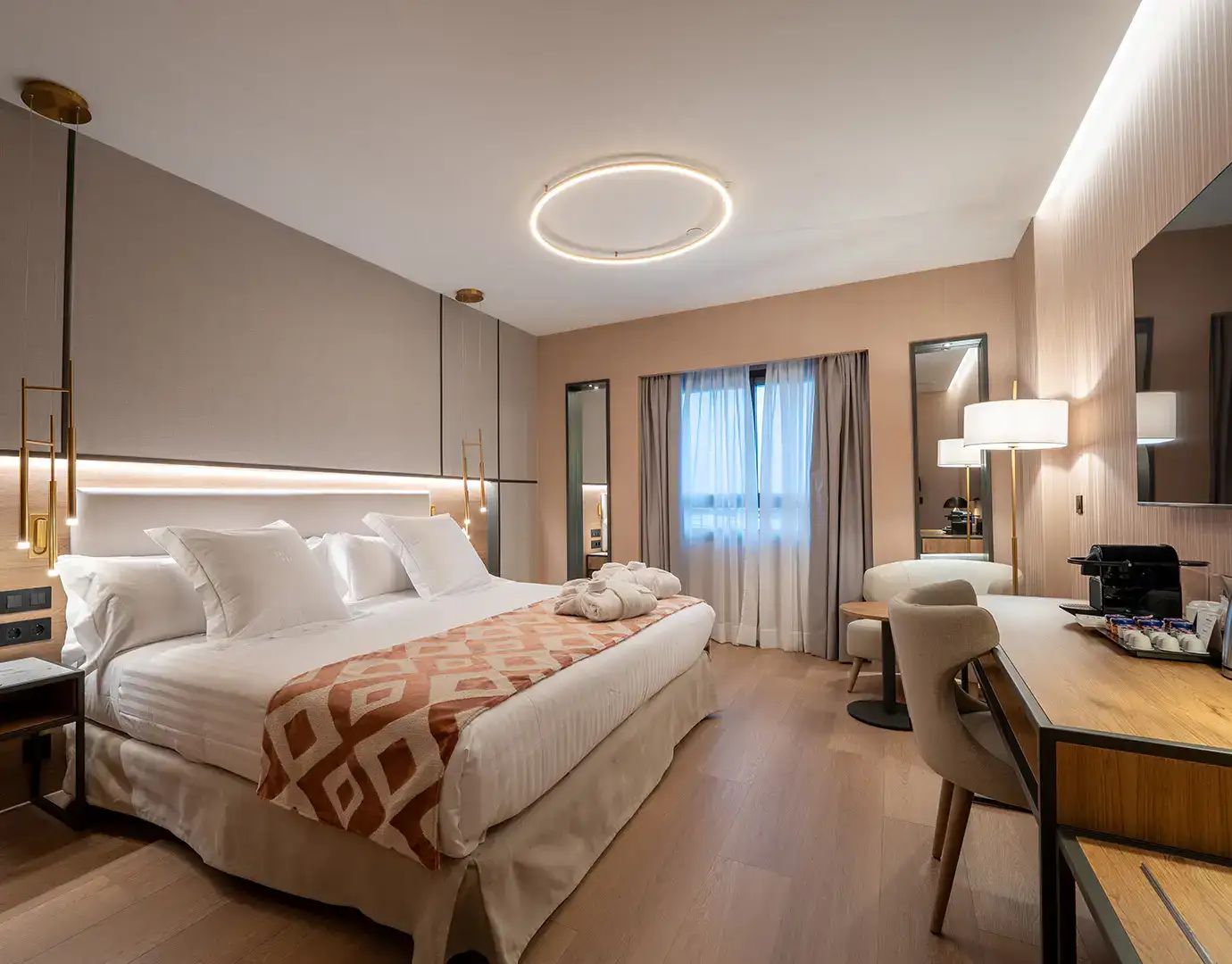 Habitaci&oacute;n de hotel moderna con cama grande, escritorio, l&aacute;mpara y ventana.