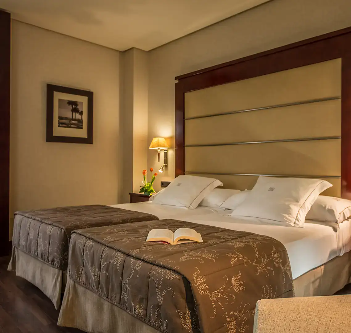 Habitaci&oacute;n de hotel acogedora con dos camas, libro sobre una y l&aacute;mpara encendida.