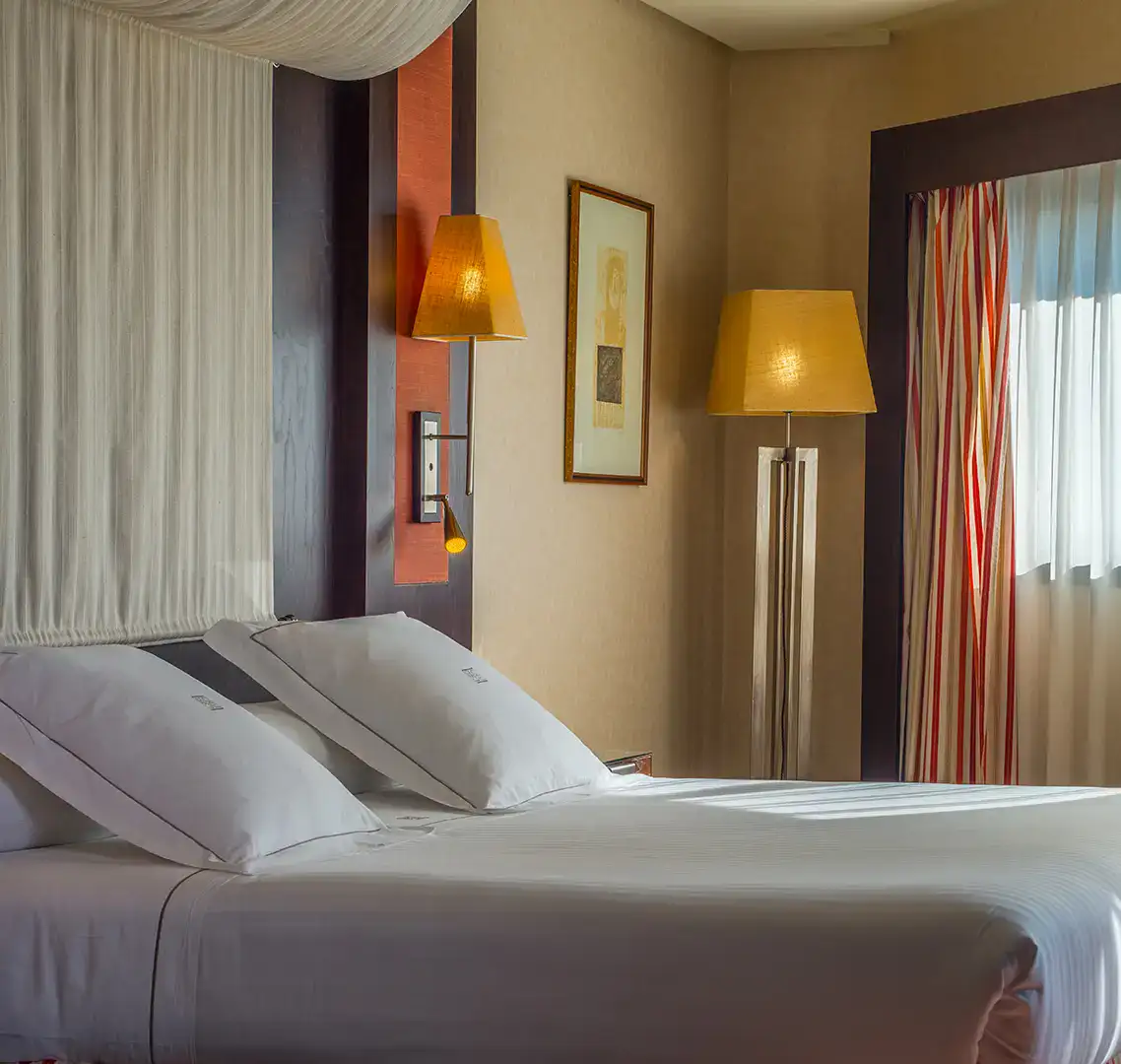 Cama con almohadas en habitaci&oacute;n iluminada con l&aacute;mparas y cortinas a rayas.