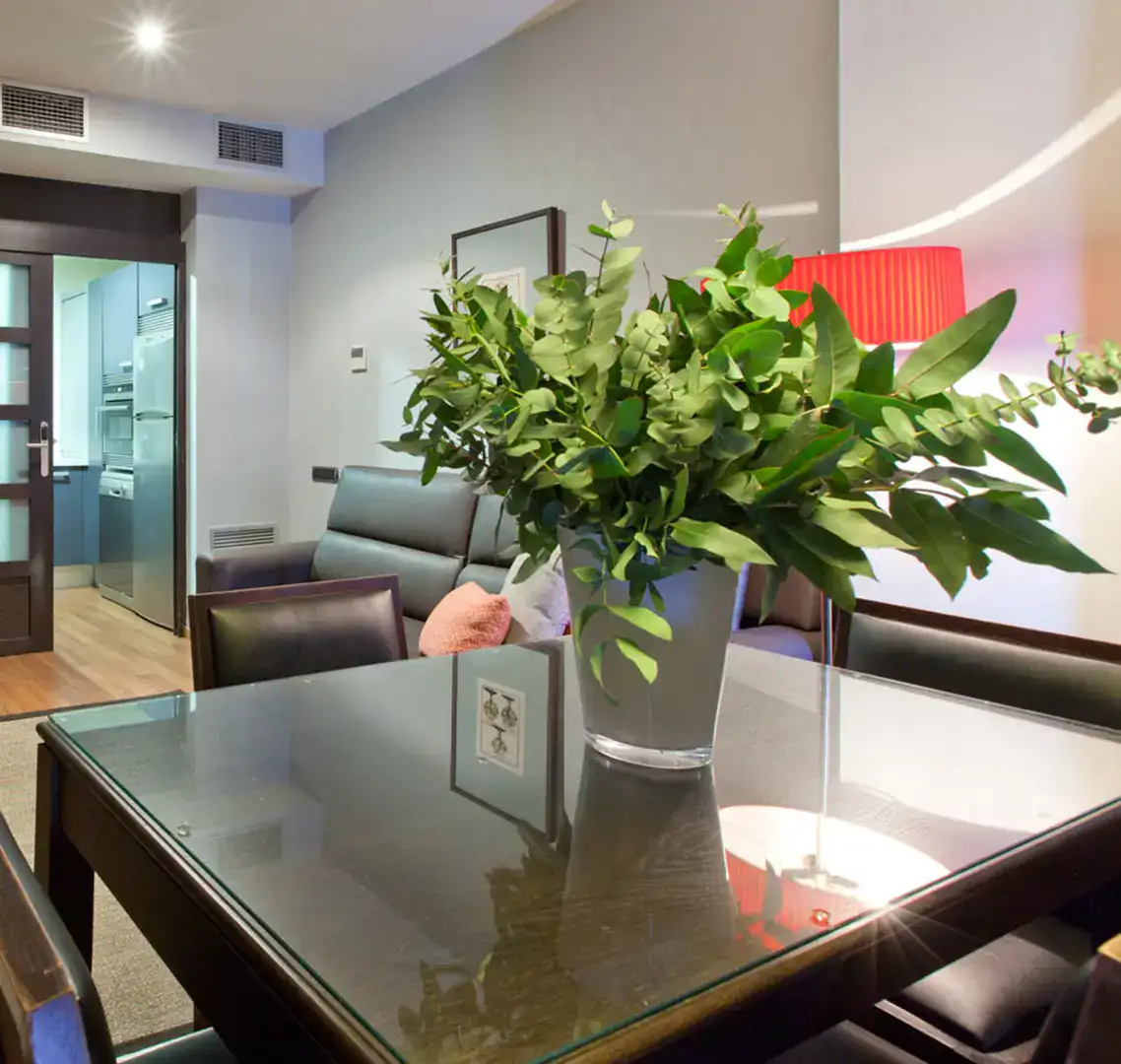 Sala moderna con mesa, jarr&oacute;n de hojas y l&aacute;mpara roja.