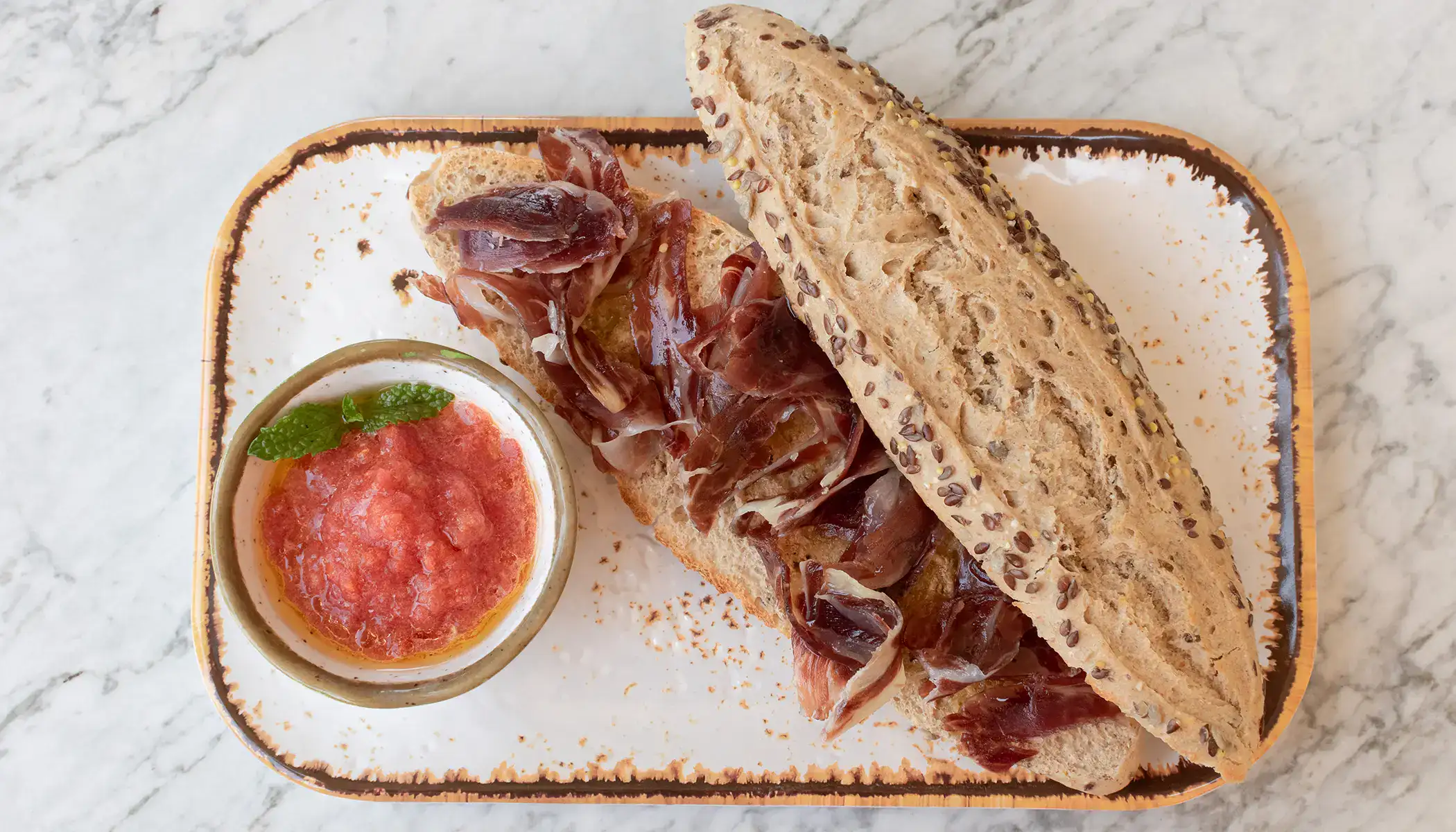 Bocadillo de jam&oacute;n sobre plato con taz&oacute;n de tomate triturado y menta.