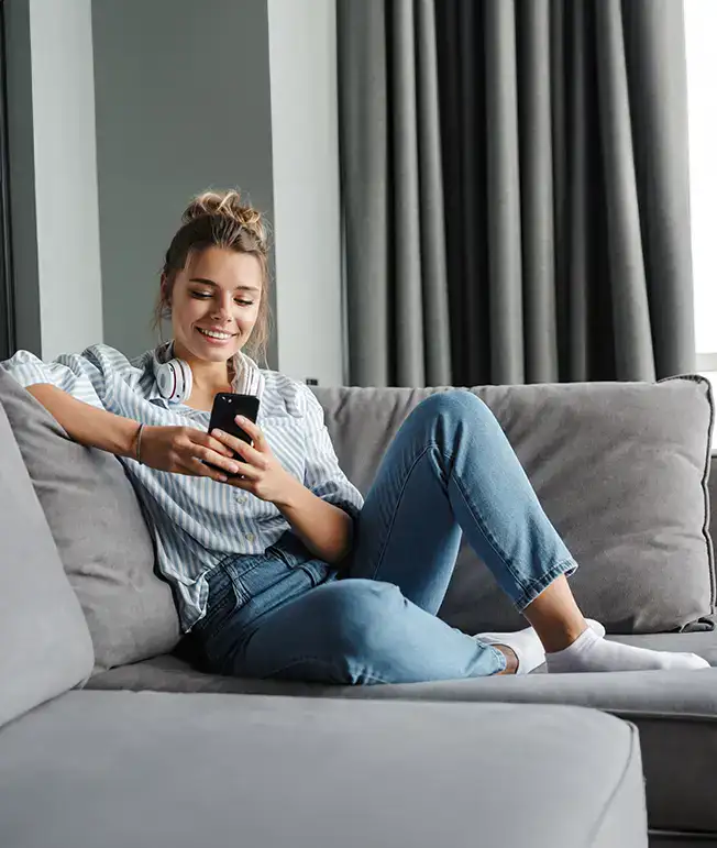Mujer sonriente en el sof&aacute; usando un celular y auriculares en su cuello.