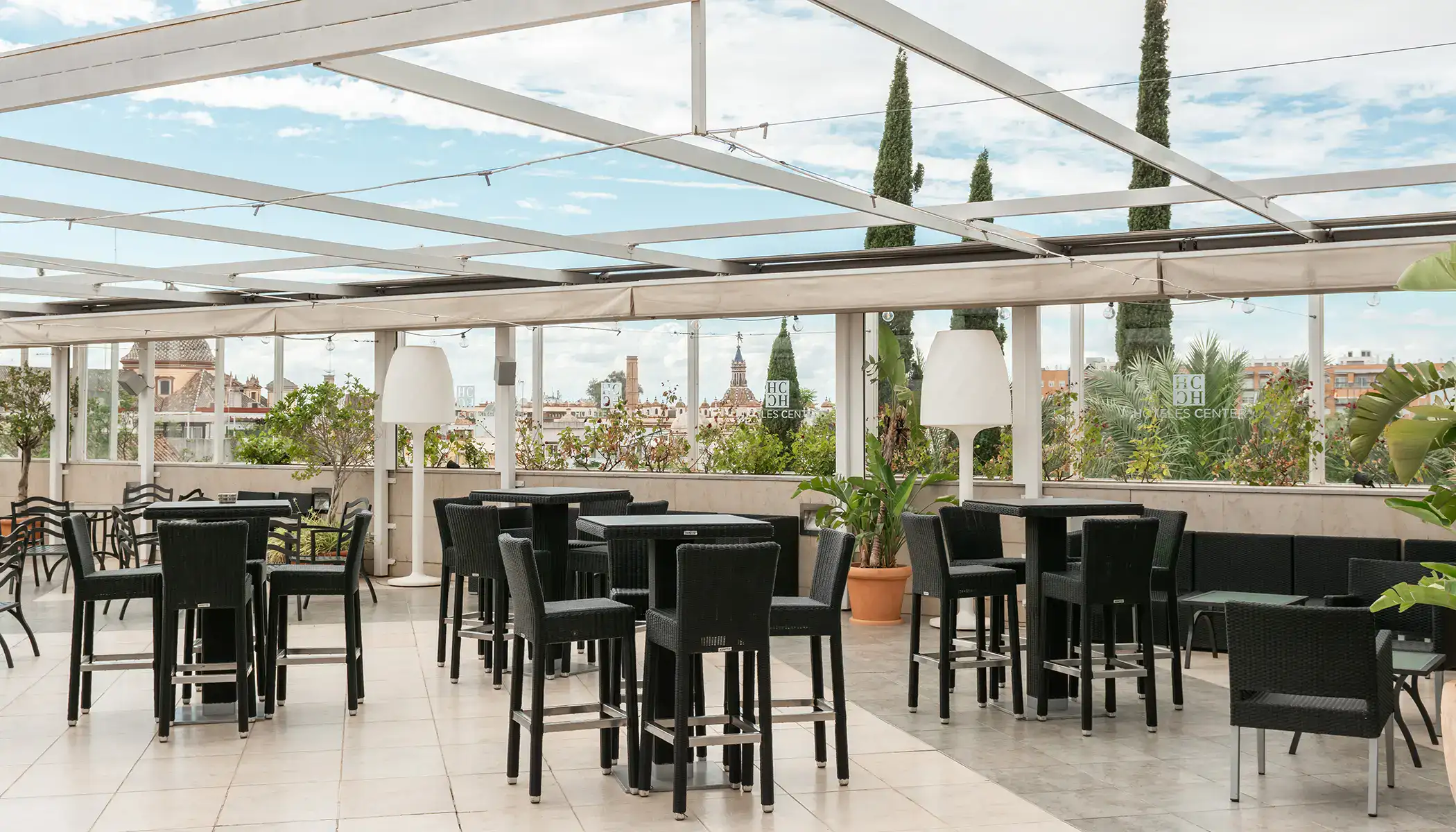 Terraza con sillas y mesas altas, rodeada de plantas y vista urbana.
