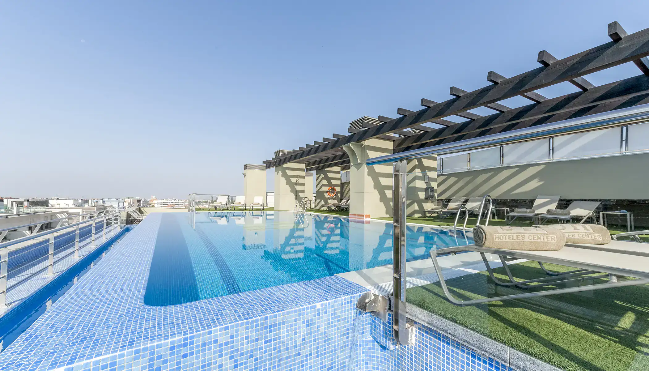 Piscina infinita en la azotea con tumbonas y p&eacute;rgolas bajo un cielo despejado.