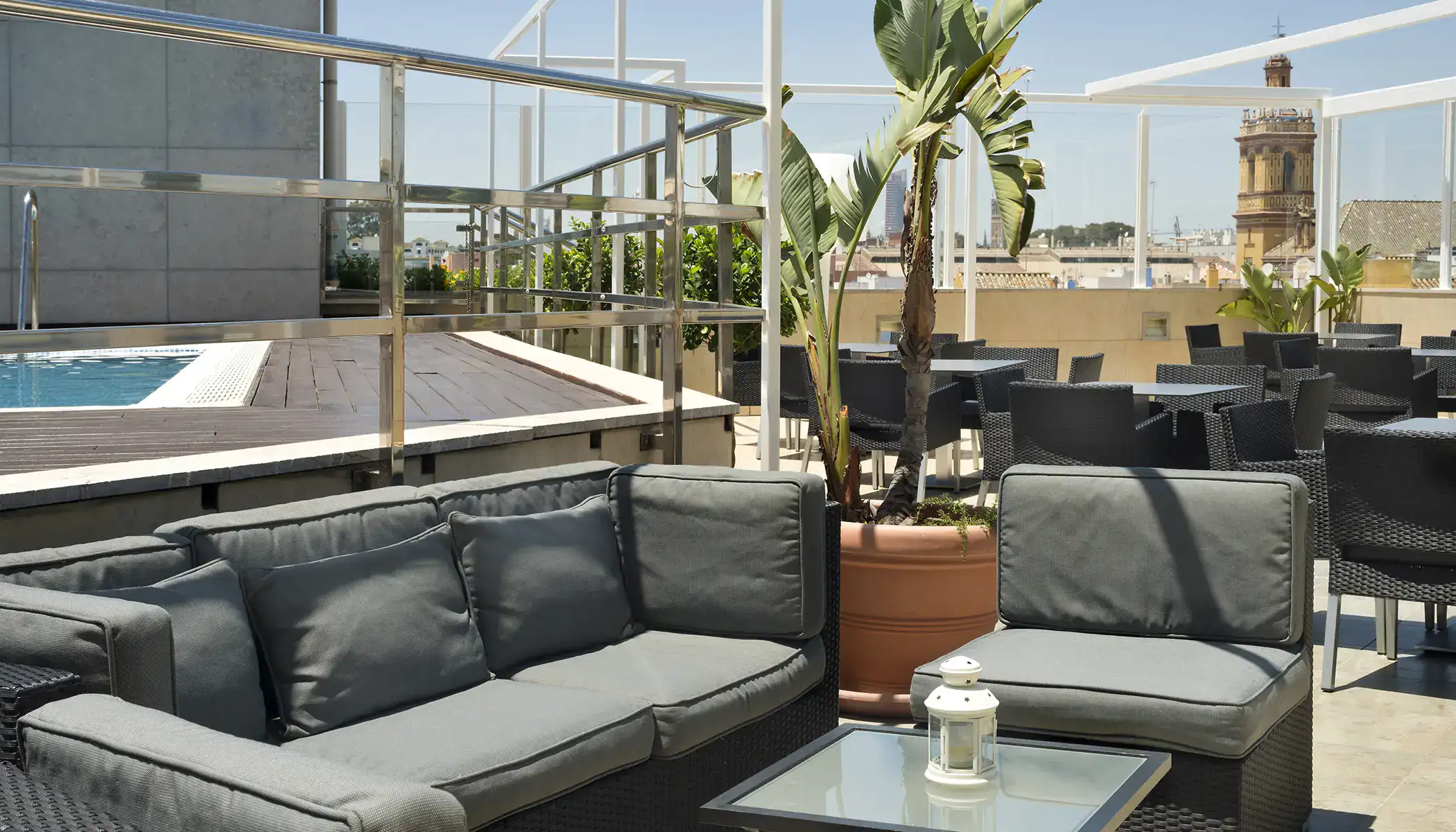 Terraza con sof&aacute;s grises, mesa de vidrio, piscina y plantas al fondo.