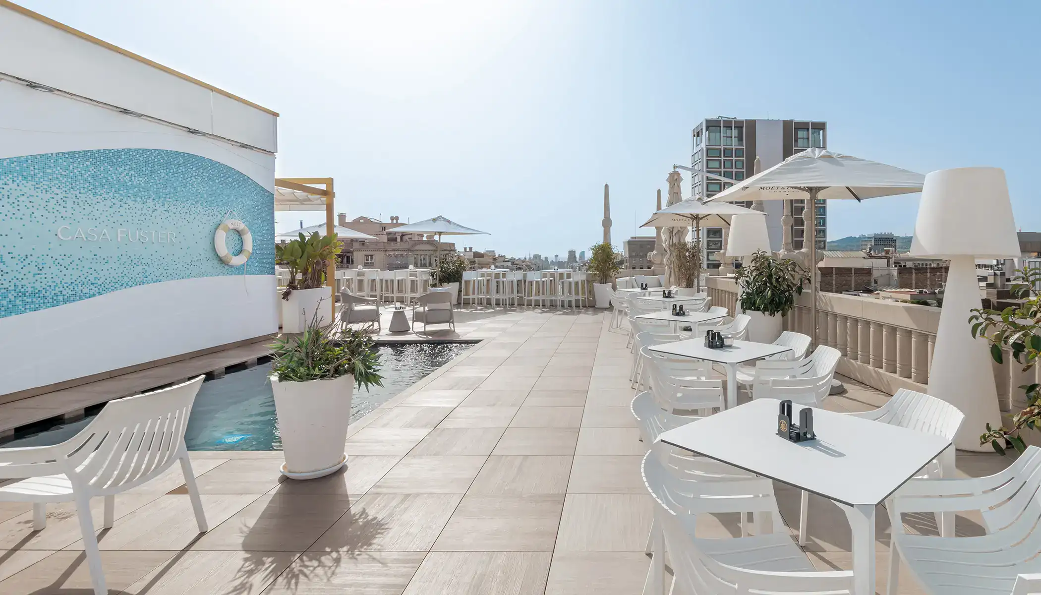 Terraza con mesas y sillas blancas junto a una piscina, d&iacute;a soleado.