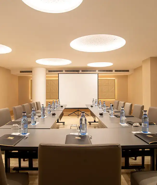 Sala de conferencias con mesa en U, proyectores y botellas de agua.