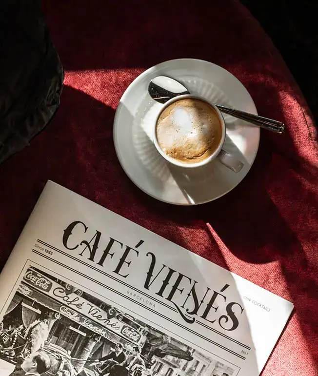Taza de caf&eacute; con revista "Caf&eacute; Vien&eacute;s" sobre una mesa con mantel rojo.