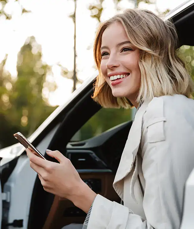 Mujer sonriendo mientras sostiene un celular al lado de un auto.