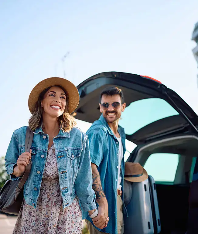Pareja sonriente de pie junto a un auto, listos para viajar bajo un cielo despejado.