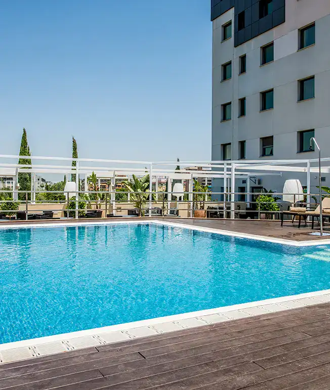 Piscina rodeada de tumbonas y plantas junto a un edificio moderno y cielo despejado.