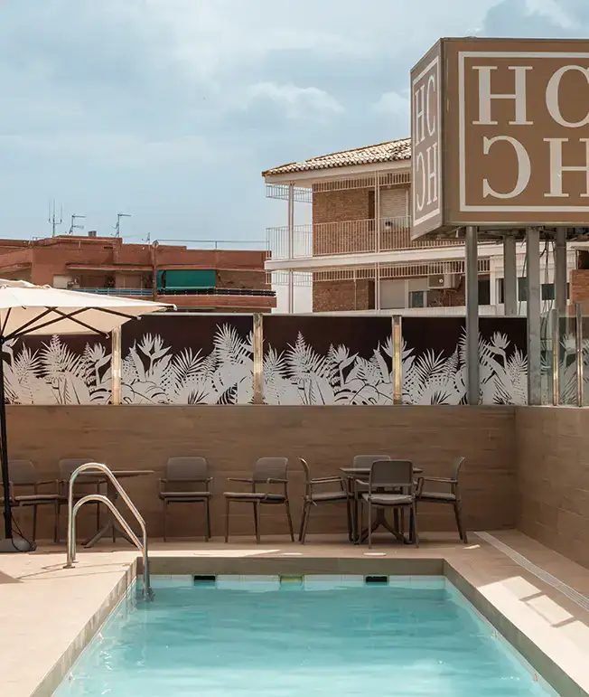 Piscina de hotel con sillas, mesas y mural de hojas al fondo.