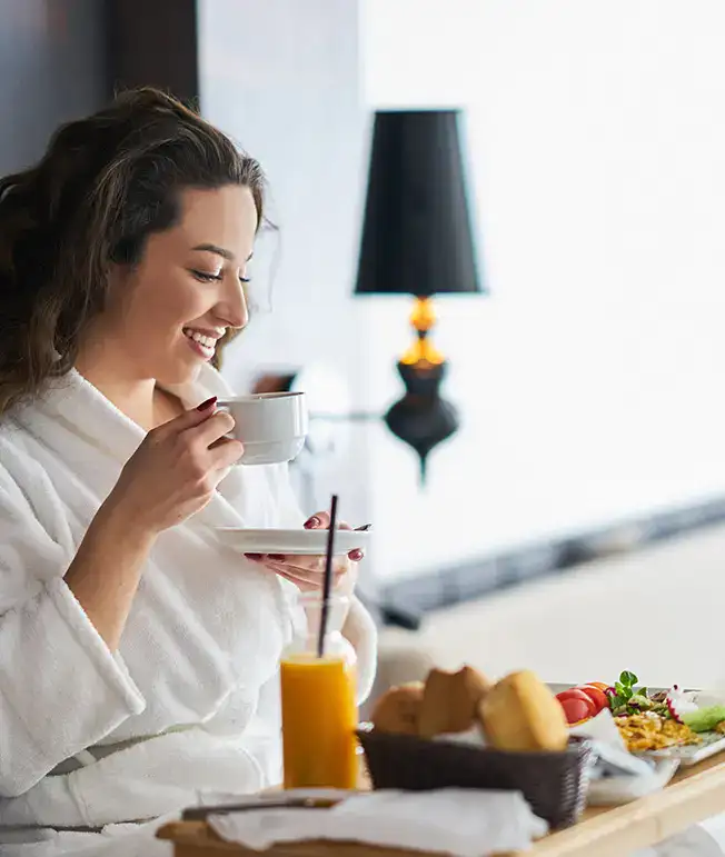 Mujer en bata tomando caf&eacute; con desayuno en una habitaci&oacute;n iluminada.