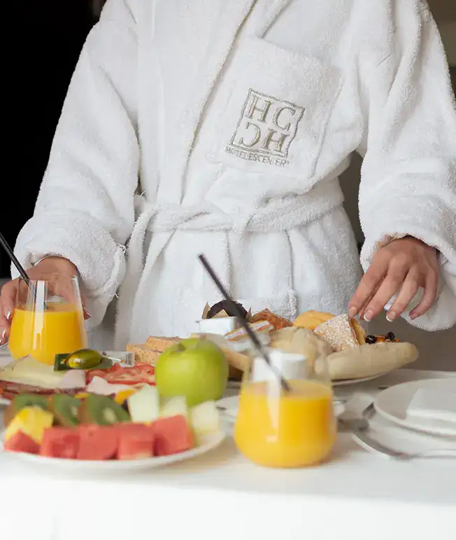 Persona en bata blanca sirviendo desayuno con zumo y fruta.