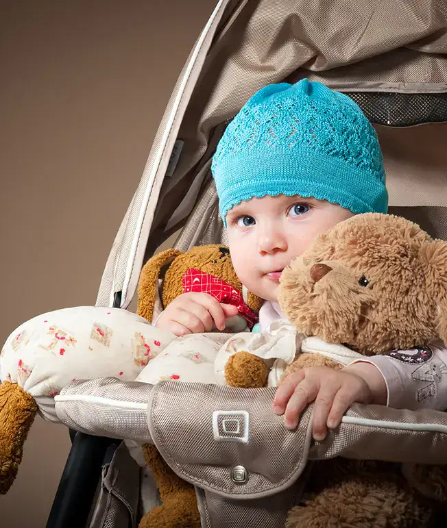 Beb&eacute; con gorro azul en cochecito, abrazando peluches beige y marr&oacute;n.