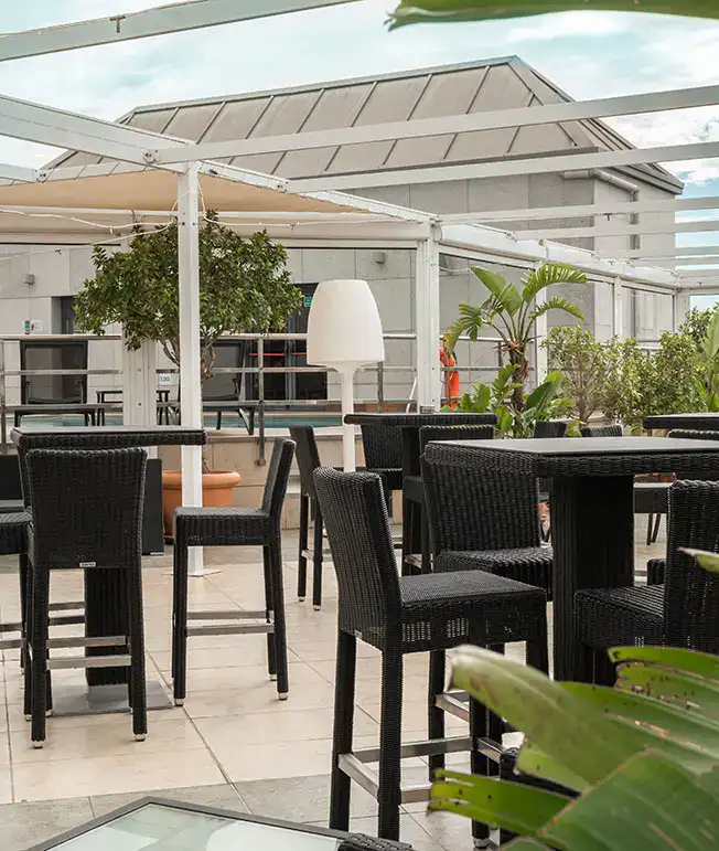 Terraza con mesas y sillas negras, plantas y estructura de p&eacute;rgola blanca.