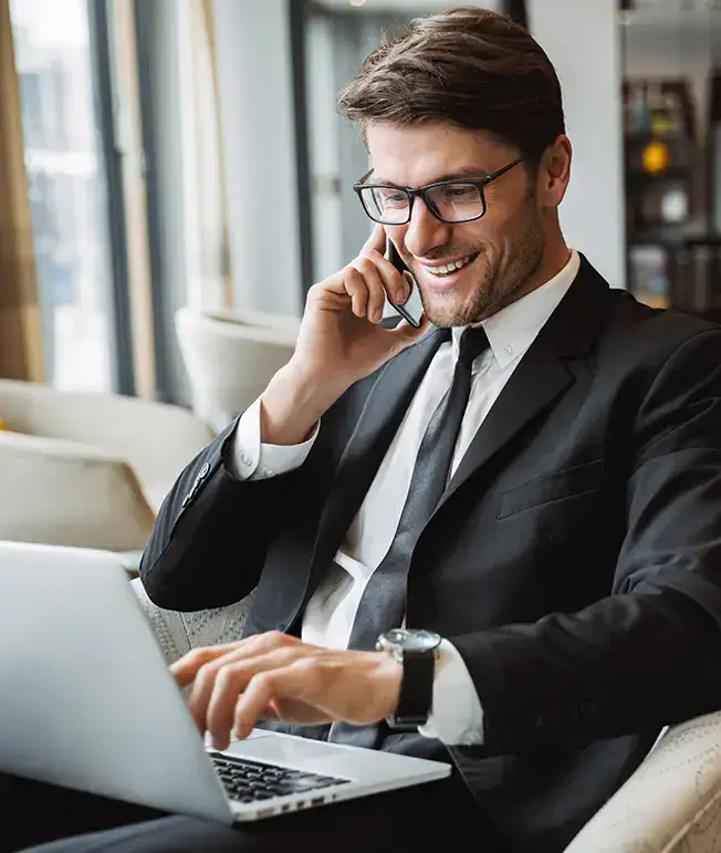 Hombre sonriente en traje, usando laptop y hablando por tel&eacute;fono.