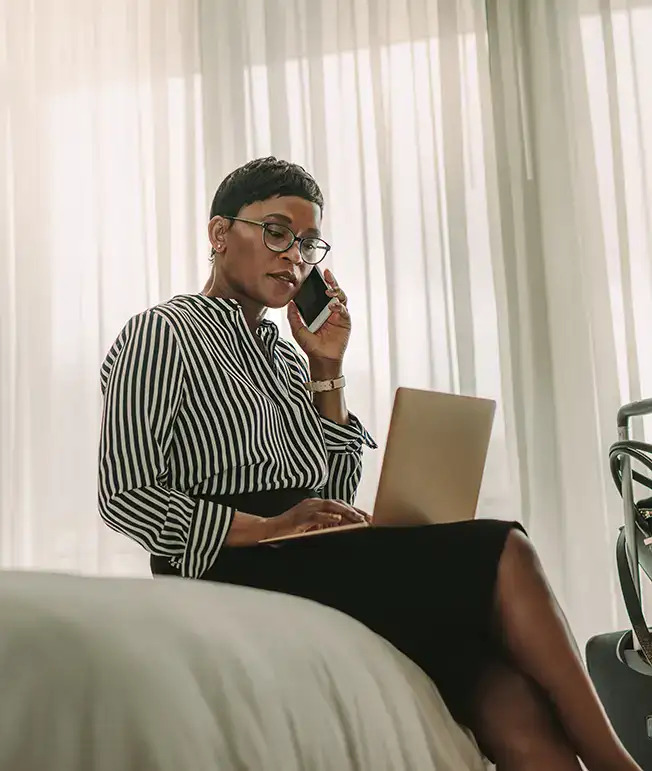 Mujer hablando por tel&eacute;fono y usando una laptop junto a una ventana.
