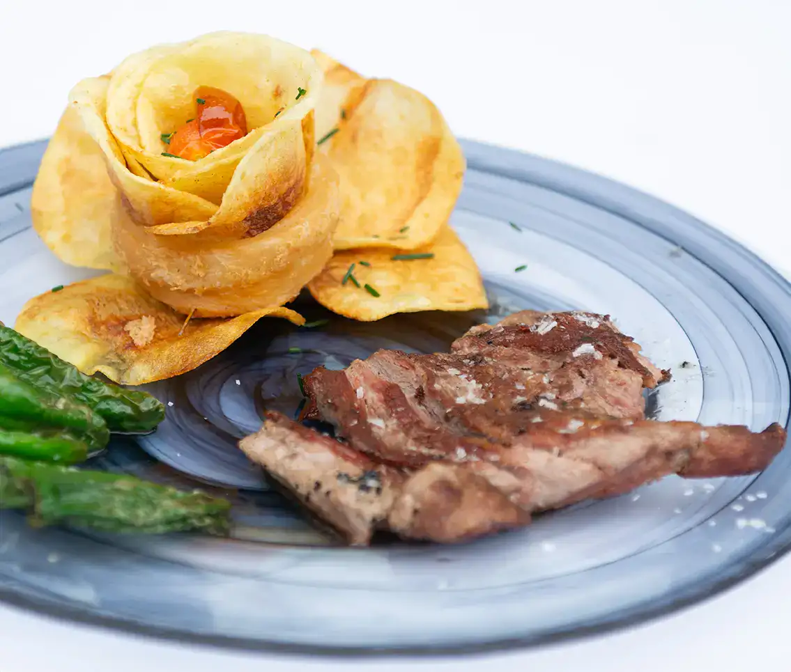 Plato con carne, flor de patata y pimientos sobre plato azul claro.