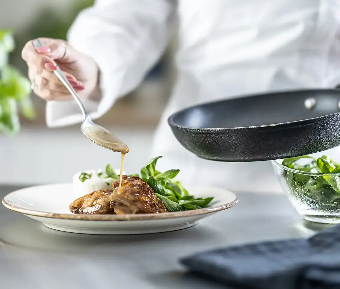 Plato servido con salsa, hojas verdes y carne, con sart&eacute;n en segundo plano.
